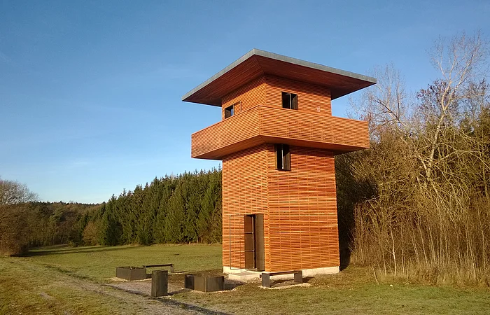 Holzbeobachtungsturm mit umlaufendem Balkon in einer Wiesenlandschaft neben Wald und kahlen Bäumen bei blauem Himmel