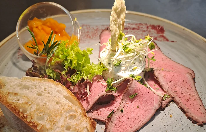 Teller mit Roastbeef, Salat, Brot, Frischkäse mit Sprossen und einem kleinen Glas mit orangener Sauce.