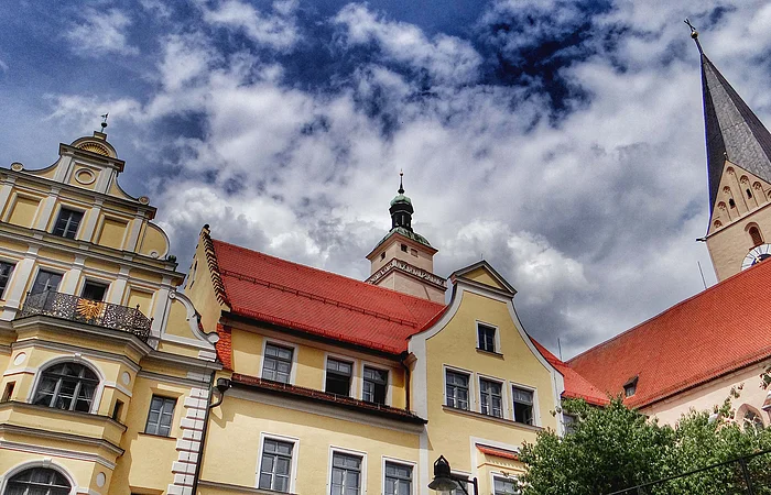 Historische gelbe Gebäude mit roten Dächern und Kirchturm unter bewölktem Himmel.