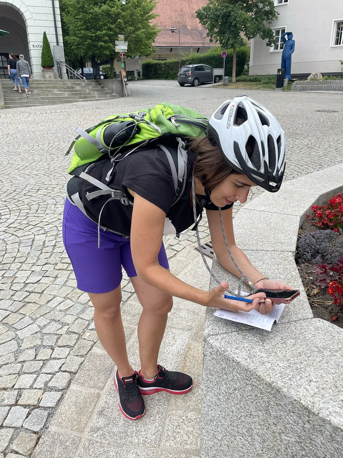 Frau mit Fahrradhelm und grünem Rucksack lehnt sich auf Steinmauer und schaut auf Handy mit Zettel und Stift.