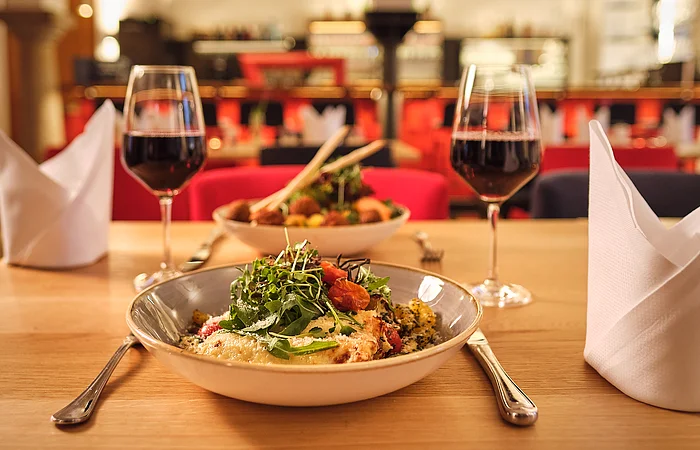 Tisch mit zwei Tellern, zwei Gläsern Rotwein, Besteck und zwei gefalteten weißen Servietten in einem Restaurant.