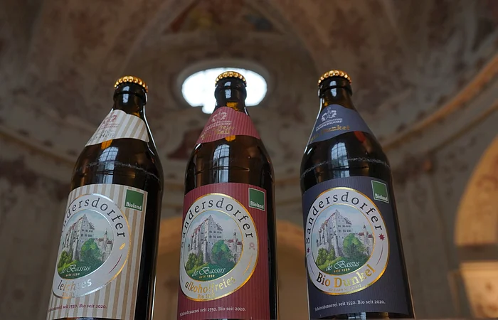 Drei Bierflaschen mit Etiketten der Marke Sandersdorfer, im Hintergrund eine verzierte Decke.