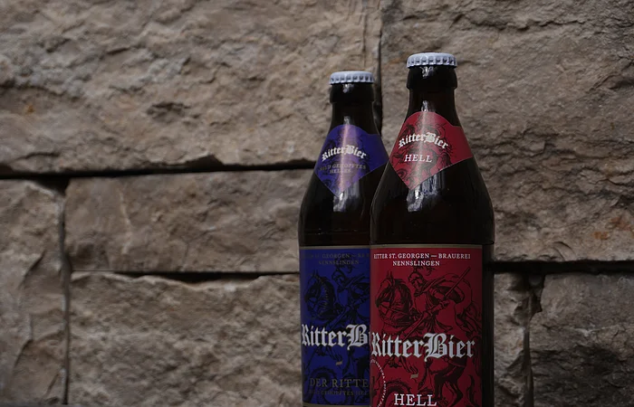 Zwei Bierflaschen mit Etiketten "RitterBier" vor einer Steinwand. Eine Flasche hat ein rotes, die andere ein blaues Etikett.