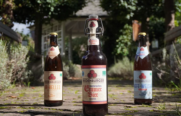 Drei Bierflaschen stehen auf einem gepflasterten Weg im Freien, umgeben von Pflanzen und Bäumen.