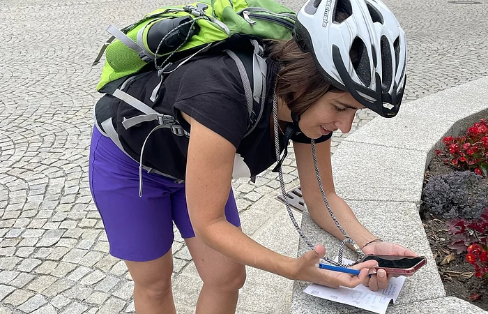 Frau mit Fahrradhelm und grünem Rucksack lehnt sich auf Steinmauer und schaut auf Handy mit Zettel und Stift.