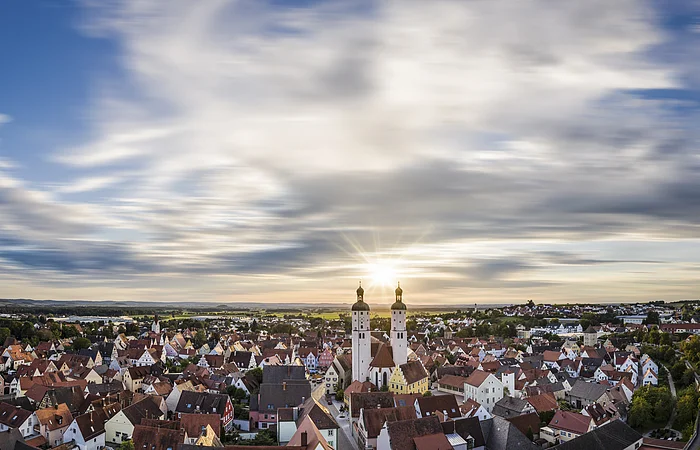 Panoramablick auf Stadt mit roten Dächern und zwei Kirchtürmen bei Sonnenuntergang unter bewölktem Himmel