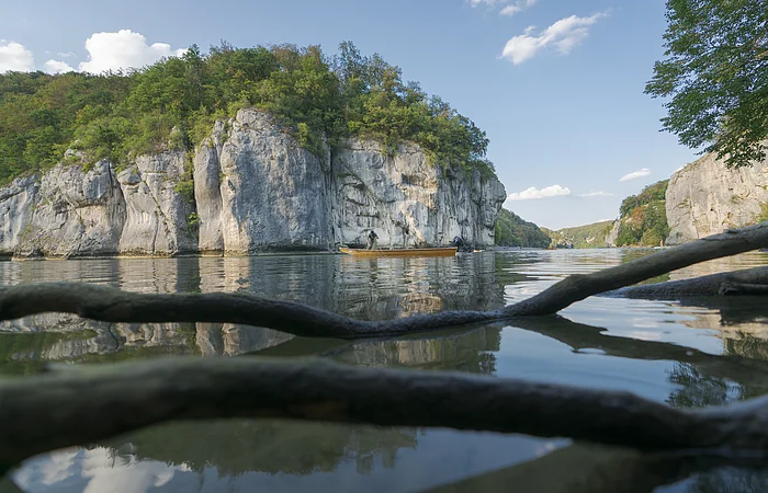 Fluss mit Felsen und Bäumen, Boot mit zwei Personen, Äste im Vordergrund im Wasser sichtbar