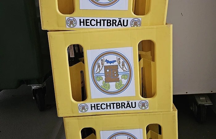 Drei gelbe Getränkekisten mit dem Aufdruck „HECHTBRÄU“ und einem runden Logo mit HB und Hecht, gestapelt auf grauem Boden.