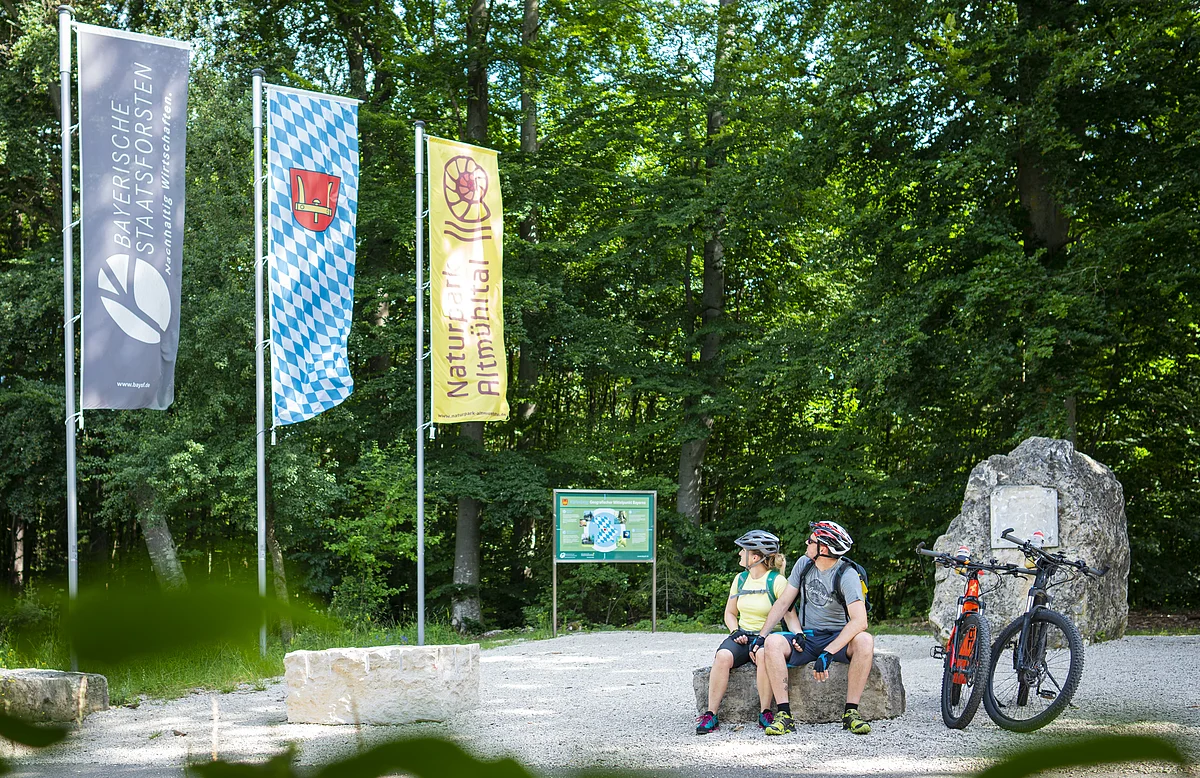 Zwei Radfahrer mit Helm sitzen auf Steinbank im Wald neben zwei Mountainbikes und drei Fahnen.