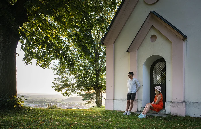 Mann lehnt an Hauswand, Frau sitzt in Türbogen, große Bäume, Dorf im Hintergrund bei Tageslicht.