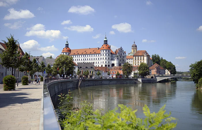 Flussufer mit Promenade und Bäumen, im Hintergrund Schloss mit Türmen und Brücke bei klarem Himmel.