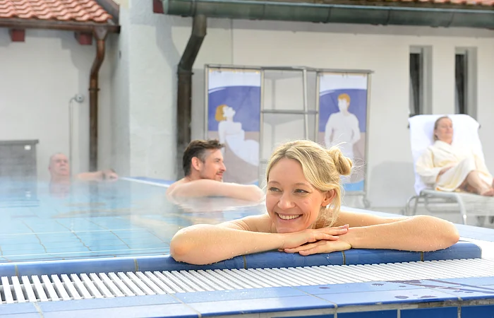 Frau lehnt entspannt am Beckenrand eines Hallenbads, im Hintergrund zwei Männer im Wasser und eine Frau auf Liegestuhl.