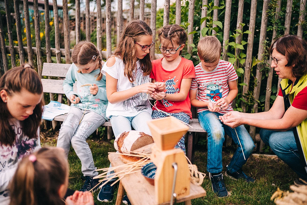 Kinder und Erwachsene sitzen auf einer Bank im Garten und bearbeiten Getreide mit einer Mühle aus Holz.