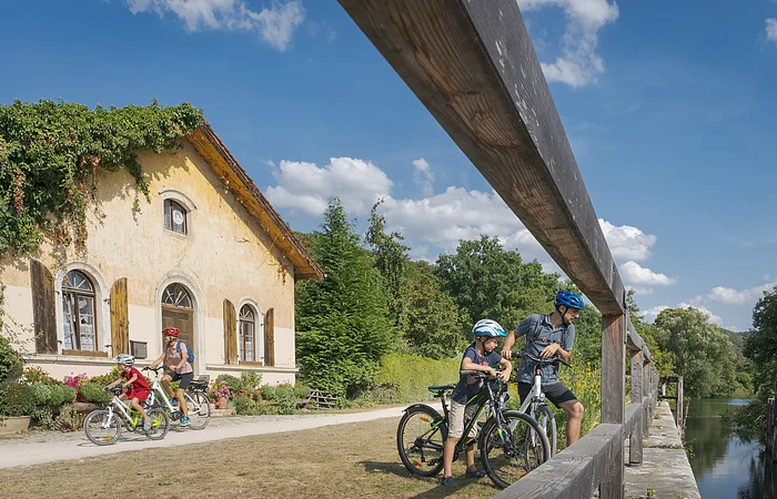 Familie mit vier Personen und Fahrrädern neben einem alten Haus und einem Fluss unter blauem Himmel.