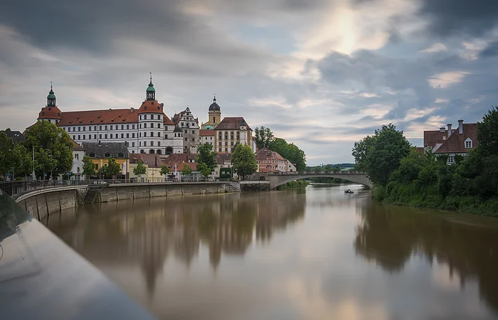 Fluss mit Brücke und historischen Gebäuden im Hintergrund, bewölkter Himmel.