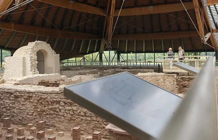Überdachte archäologische Ausgrabung mit zwei Personen auf einem Holzsteg und Informationstafel im Vordergrund.