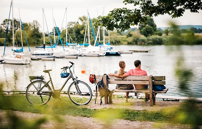 Paar sitzt auf Holzbank am See, Fahrrad mit Helm lehnt daneben, Segelboote im Hintergrund sichtbar.