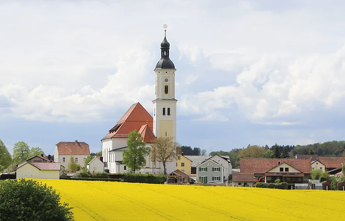 Dorf mit Kirche, Häusern und gelbem Rapsfeld unter bewölktem Himmel.