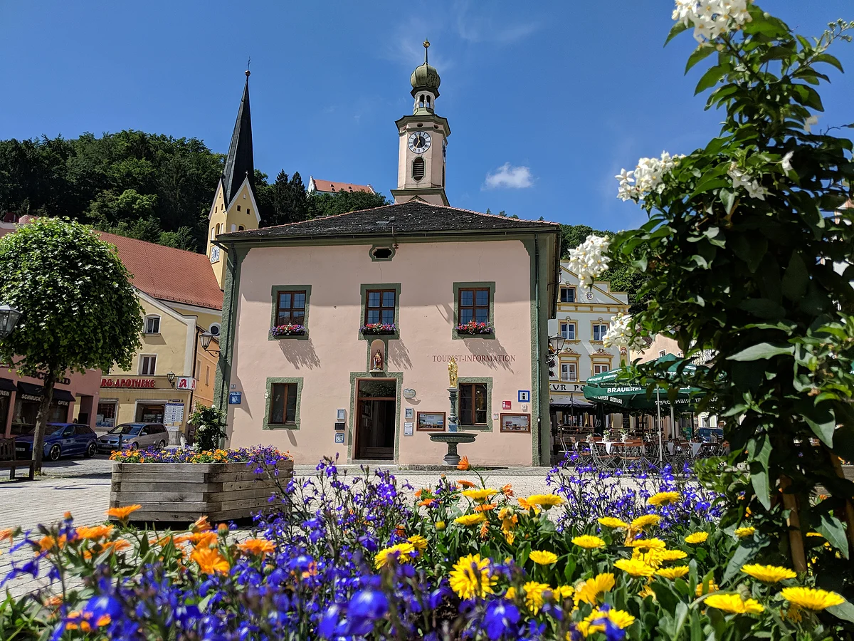 Gebäude mit Turmuhr und Touristeninformation, davor bunte Blumen und blauer Himmel an einem sonnigen Tag.