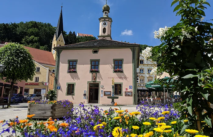 Gebäude mit Turmuhr und Touristeninformation, davor bunte Blumen und blauer Himmel an einem sonnigen Tag.