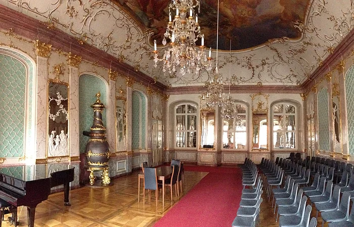 Prunkvoller Saal mit Kronleuchtern, roten Teppich, Klavier, Stühlen und goldverzierten Wänden und Decke.