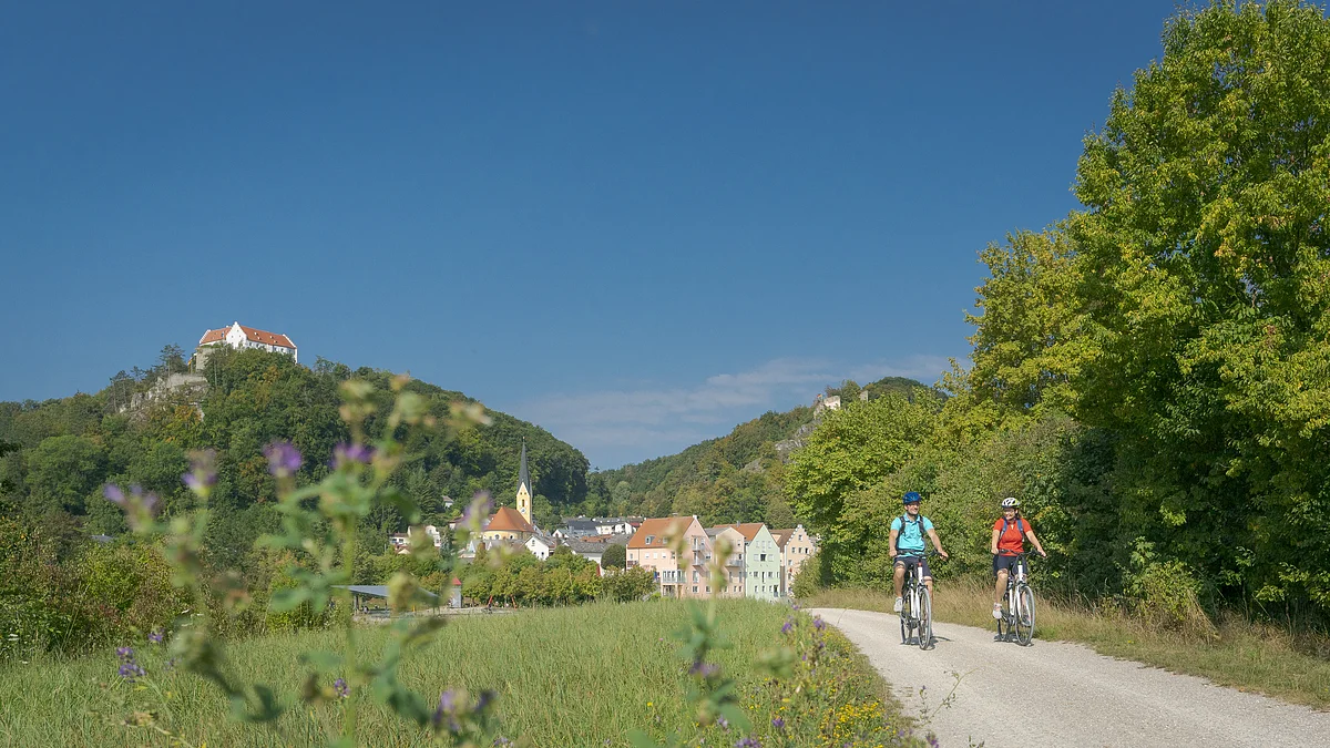 Zwei Radfahrer auf einem Weg neben grünen Bäumen mit Dorf und Burg auf Hügeln im Hintergrund bei blauem Himmel.