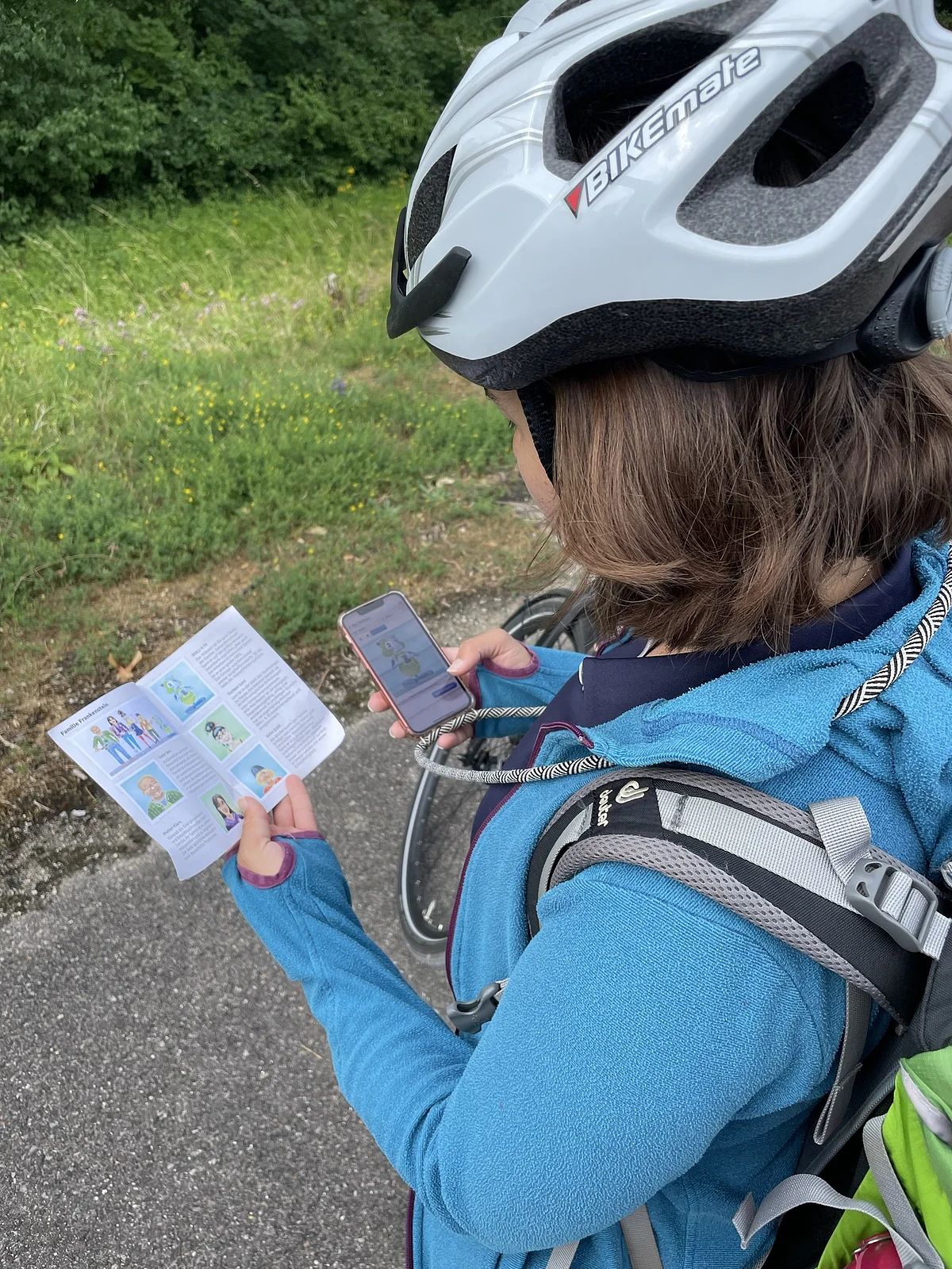 Person mit Fahrradhelm und Rucksack hält Flyer und Smartphone, steht auf Weg neben Wiese mit Bäumen.