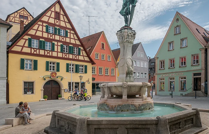 Brunnen mit Statue auf einem Platz, umgeben von Fachwerkhäusern und Menschen, unter bewölktem Himmel.