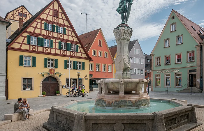 Brunnen mit Statue auf einem Platz, umgeben von Fachwerkhäusern und Menschen, unter bewölktem Himmel.