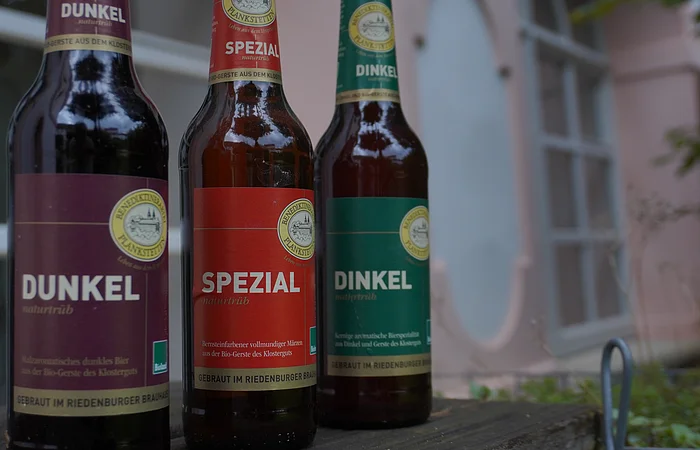 Drei Bierflaschen mit Etiketten: Dunkel, Spezial und Dinkel, auf einem Holztisch vor einem Fenster.