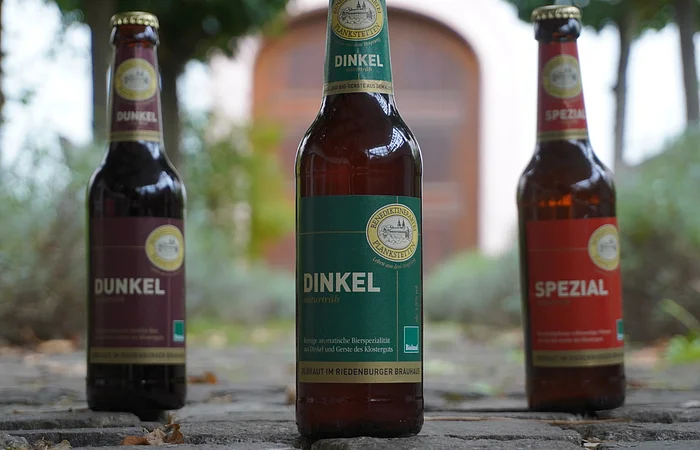 Drei Bierflaschen auf einem gepflasterten Weg, im Hintergrund unscharfe Bäume und ein Torbogen.