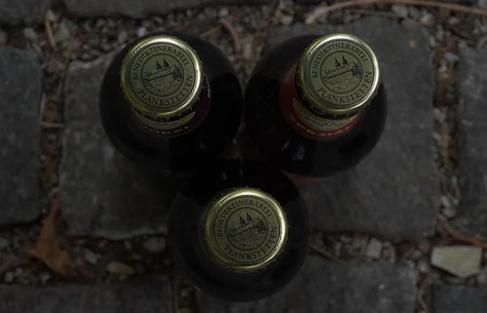 Drei Bierflaschen mit goldenen Kronkorken, auf Kopfsteinpflaster stehend, von oben fotografiert.