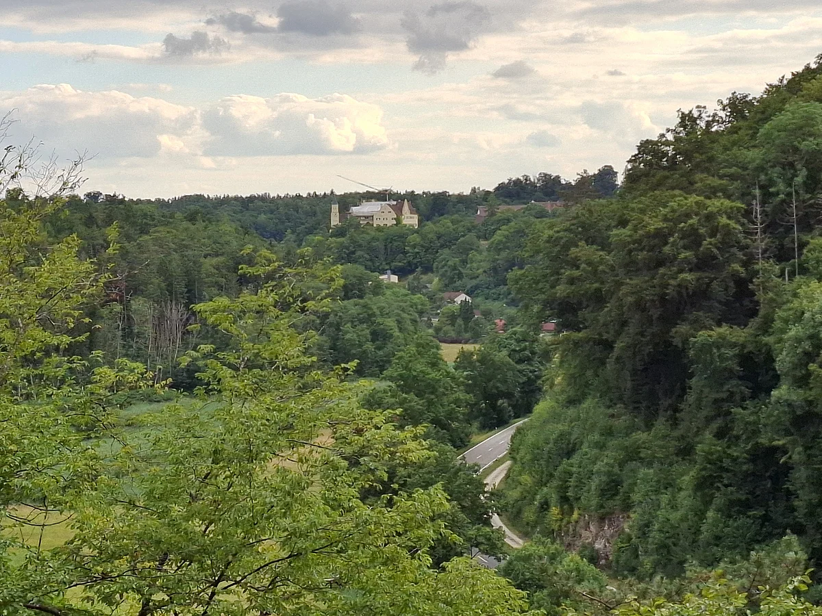 Blick auf bewaldete Hügel mit einer Straße und einem Schloss Hexenagger im Hintergrund unter bewölktem Himmel.