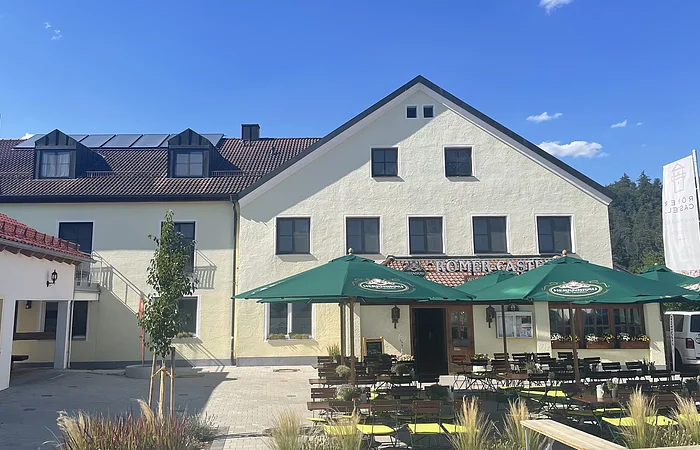 Außenansicht eines gelben Gasthofs mit grünen Sonnenschirmen und bestuhltem Biergarten bei Sonnenschein.