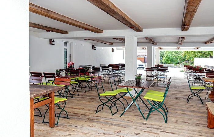 Überdachter Außenbereich mit Holzboden, Holztischen und Stühlen mit grünen Sitzkissen, weiße Wände und Decke mit Holzbalken.