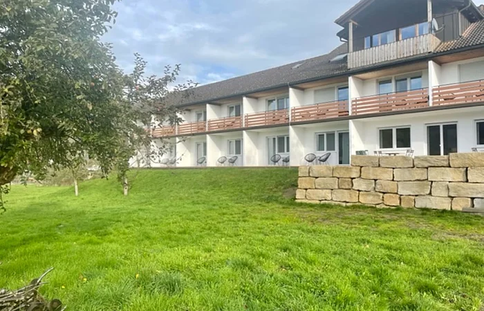 Mehrstöckiges Wohngebäude mit Balkonen, davor eine grüne Wiese und ein Baum links im Bild.