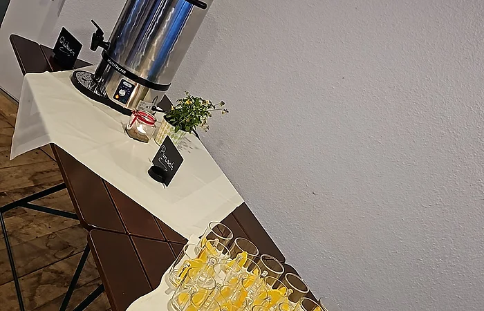 Tisch mit Gläsern und Orangenscheiben, Thermoskanne, Blumen und Kerze vor weißer Wand in Raum mit Holzboden.