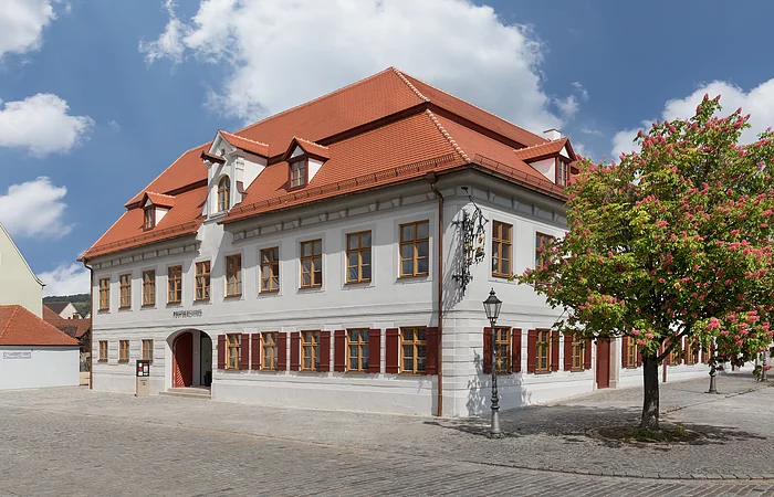 Weißes zweistöckiges Gebäude mit roten Fensterläden und Dach, blühender Baum rechts, gepflasterter Platz davor