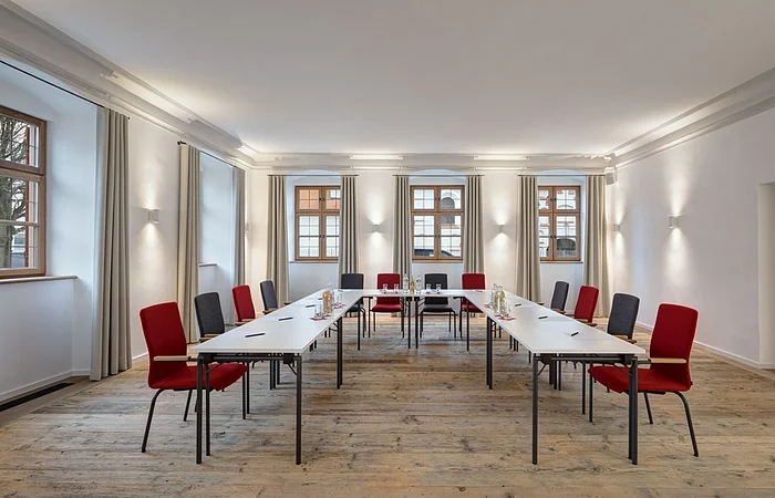 Konferenzraum mit U-förmiger Tischanordnung, roten und grauen Stühlen und Fenstern mit Vorhängen.