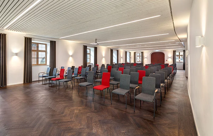 Konferenzraum mit Reihen von grauen und roten Stühlen, weißen Wänden und Holzboden.
