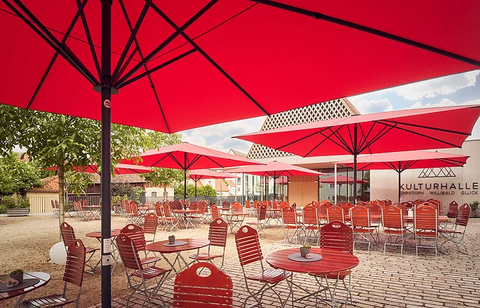 Außenbereich eines Cafés mit roten Sonnenschirmen und roten Stühlen vor einem Gebäude mit Schriftzug Kulturhalle.