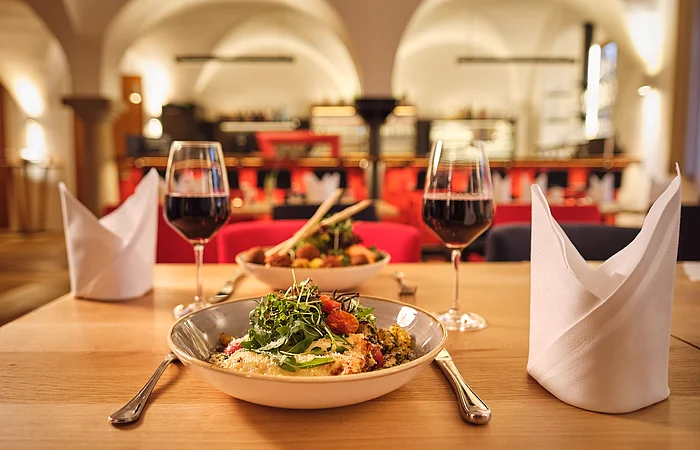 Tisch mit zwei Tellern Essen, zwei Gläsern Rotwein und gefalteten weißen Servietten in einem Restaurant mit Bogenfenstern.