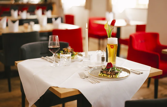Tisch in Restaurant mit weißer Tischdecke, Kerze, Glas Rotwein, Glas Bier und angerichtetem Essen.