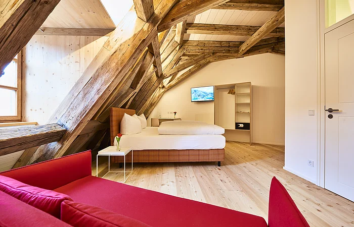 Hotelzimmer mit Holzbalken, Doppelbett, rotem Sofa, kleinem Tisch und offenem Kleiderschrank unter Dachschräge