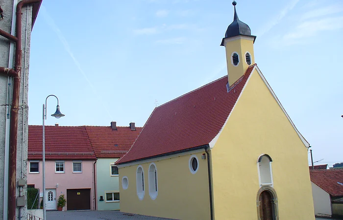 Gelbe Kapelle, die Peterskapelle in Monheim mit rotem Dach und einem Turm, umgeben von Wohnhäusern mit roten Dächern. Im Hintergrund blauer Himmel.