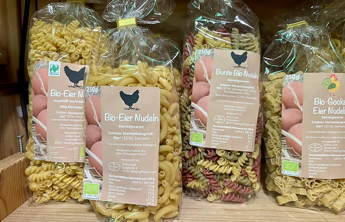 Verschiedene Packungen Bio-Eiernudeln in Plastikbeuteln auf Holzregal, Sorten und Formen sichtbar.