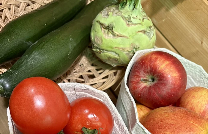 Tomaten, Äpfel, Gurken und Kohlrabi liegen auf einem Holztisch in Behältern und einem Korb.