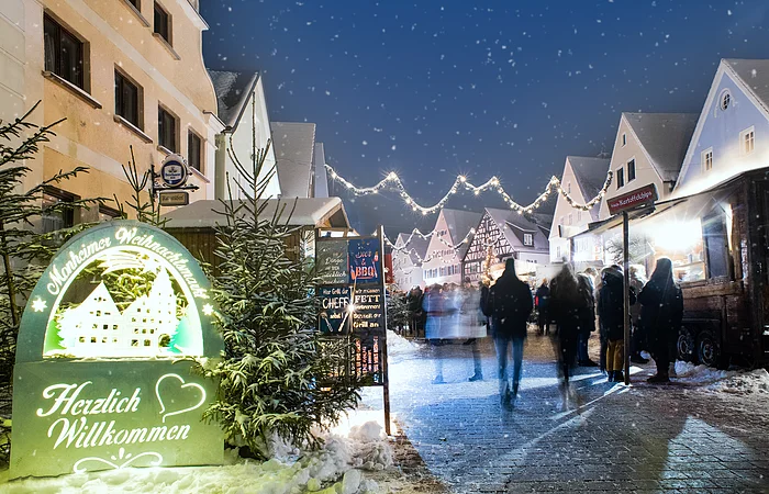 Monheimer Weihnachtsmarkt mit Verkaufsständen, Lichtern und Menschen. Ein beleuchtetes Schild links im Bild mit dem Text: "Monheimer Weihnachtsmarkt - Herzlich Willkommen" umgeben von mehreren Tannenbäumen. Schnee liegt auf dem gepflastertem Platz.