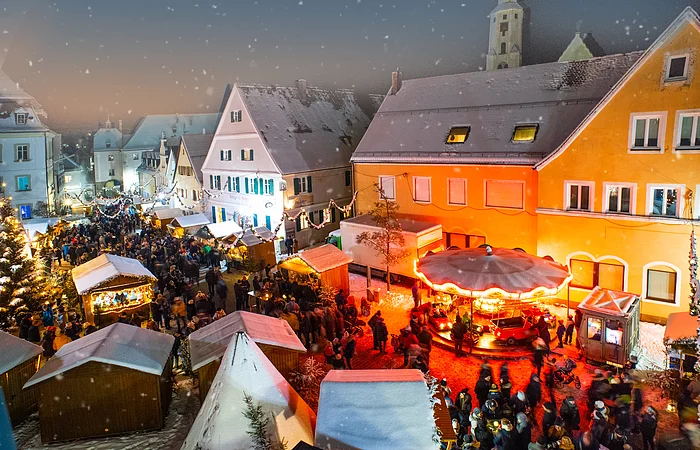 Der Monheimer Weihnachtsmarkt mit zahlreichen Besuchern, festlich beleuchteten Ständen und Kinderkarussell bei Schnee in historischen Altstadt von Monheim.