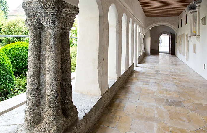 Blick in einen langen Kreuzgang mit Säulen und Rundbögen links im Bild, dahinter grüne Büsche. Rechts im Bild weiße Wand mit einer Ausfräsung in der Wand und Steinplatten mit Text angebracht. Bodenbelag aus hellen alten Steinen.